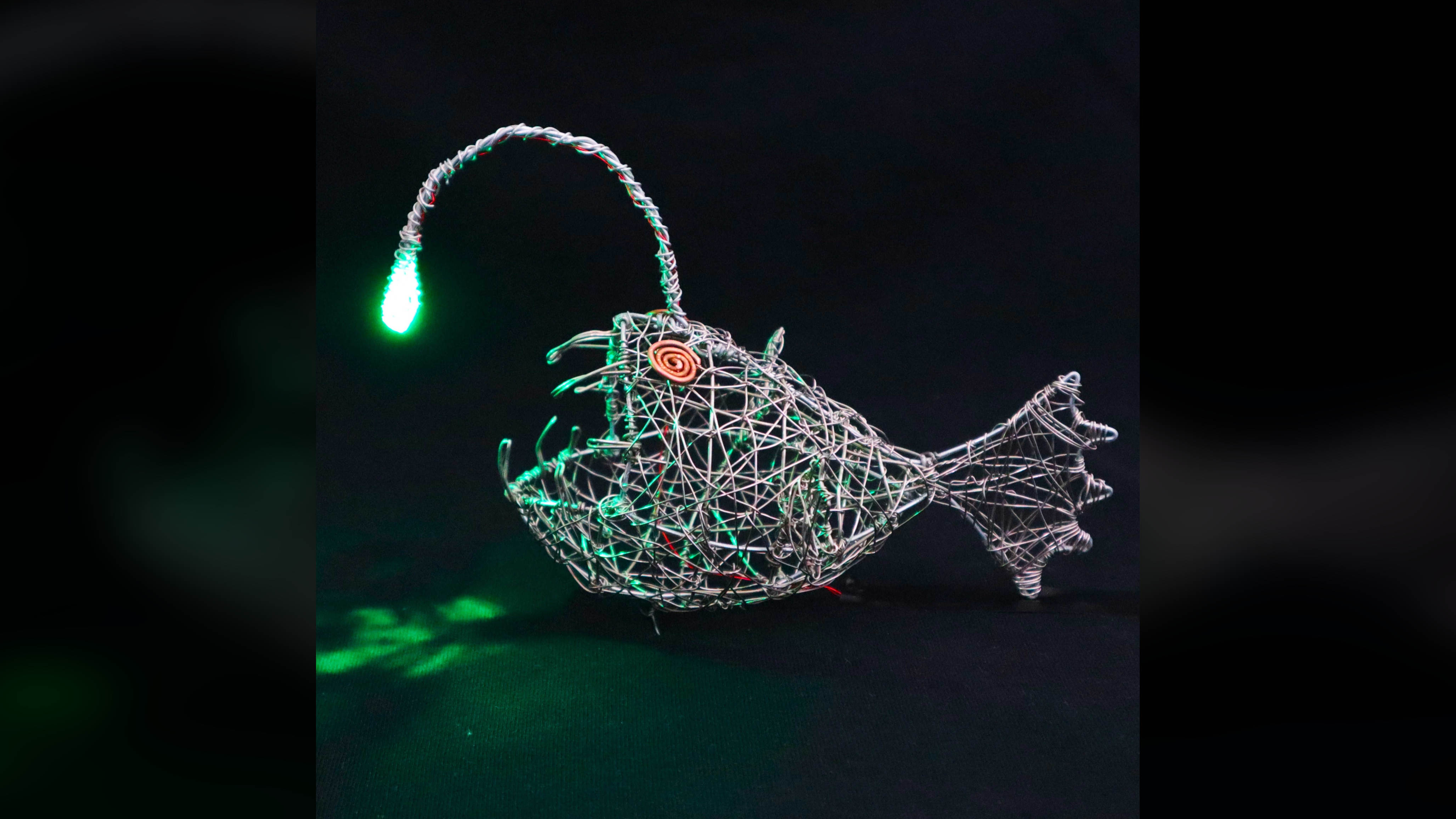 Side view of the wire anglerfish sculpture illuminated by its green LED.