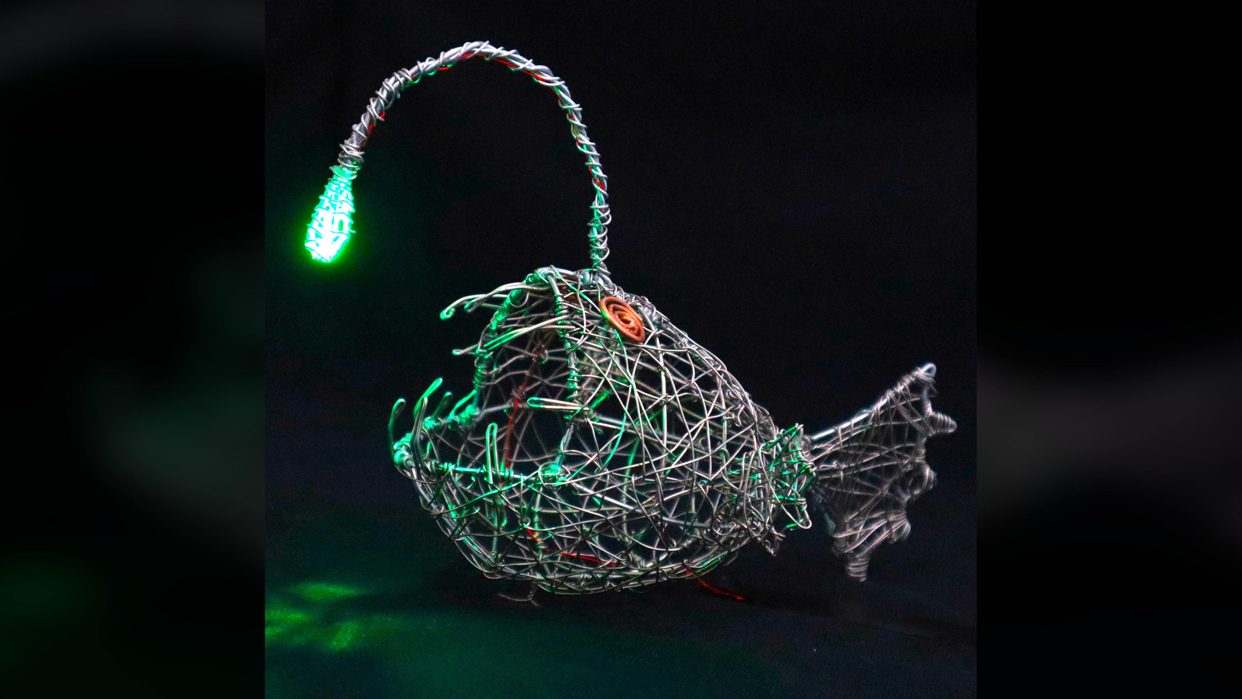 Side view of the wire anglerfish sculpture illuminated by its green LED.
